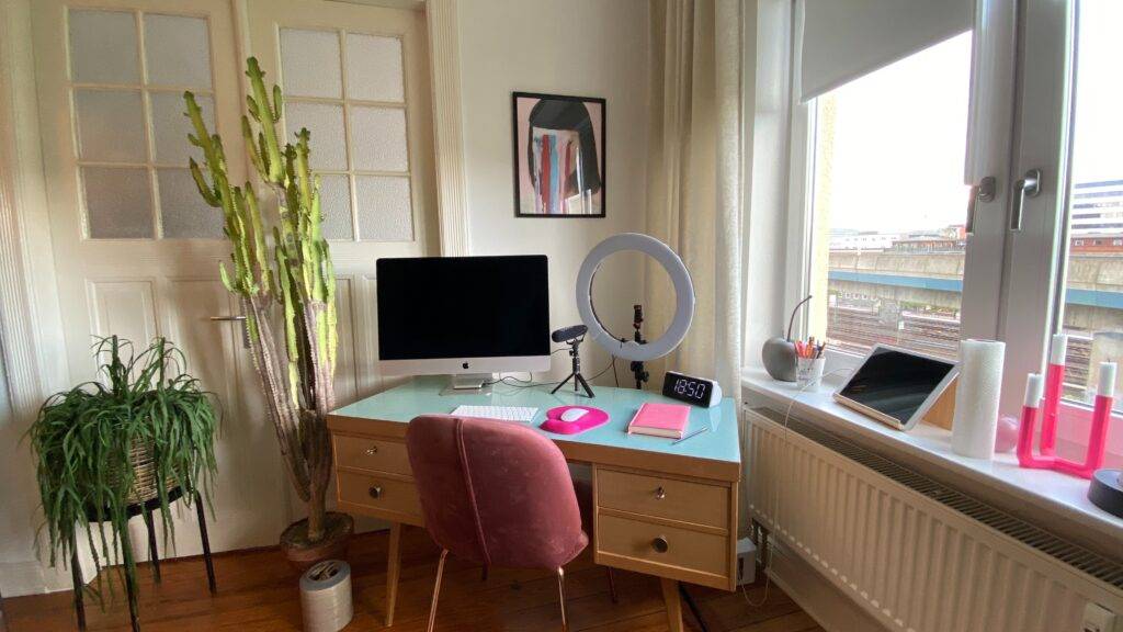 Medium writing space, desk with mac desktop and ring light, microphone, alarm clock, and journal. art on the wall, view of buildings outside.